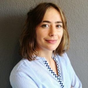 Hübsche junge Frau mit braunen Haaren und blauer Bluse, lächelnd vor grauer Wand.