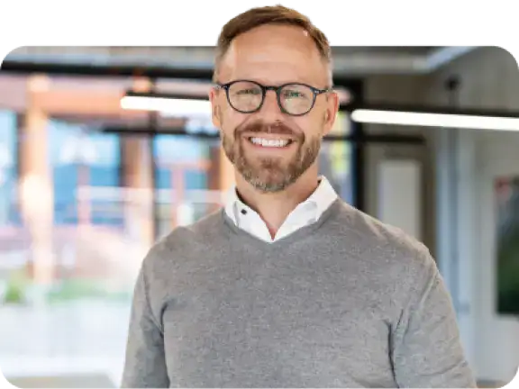Geschäftsmann mit Brille und grauem Pullover in einem hellen, modernen Büro.
