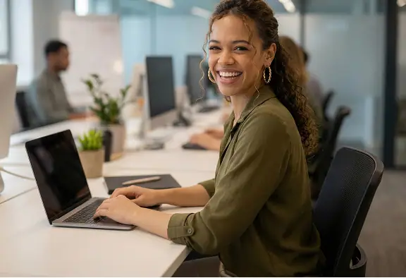Frau arbeitet lächelnd an einem Laptop im modernen Büro, umgeben von Kollegen und Pflanzen.