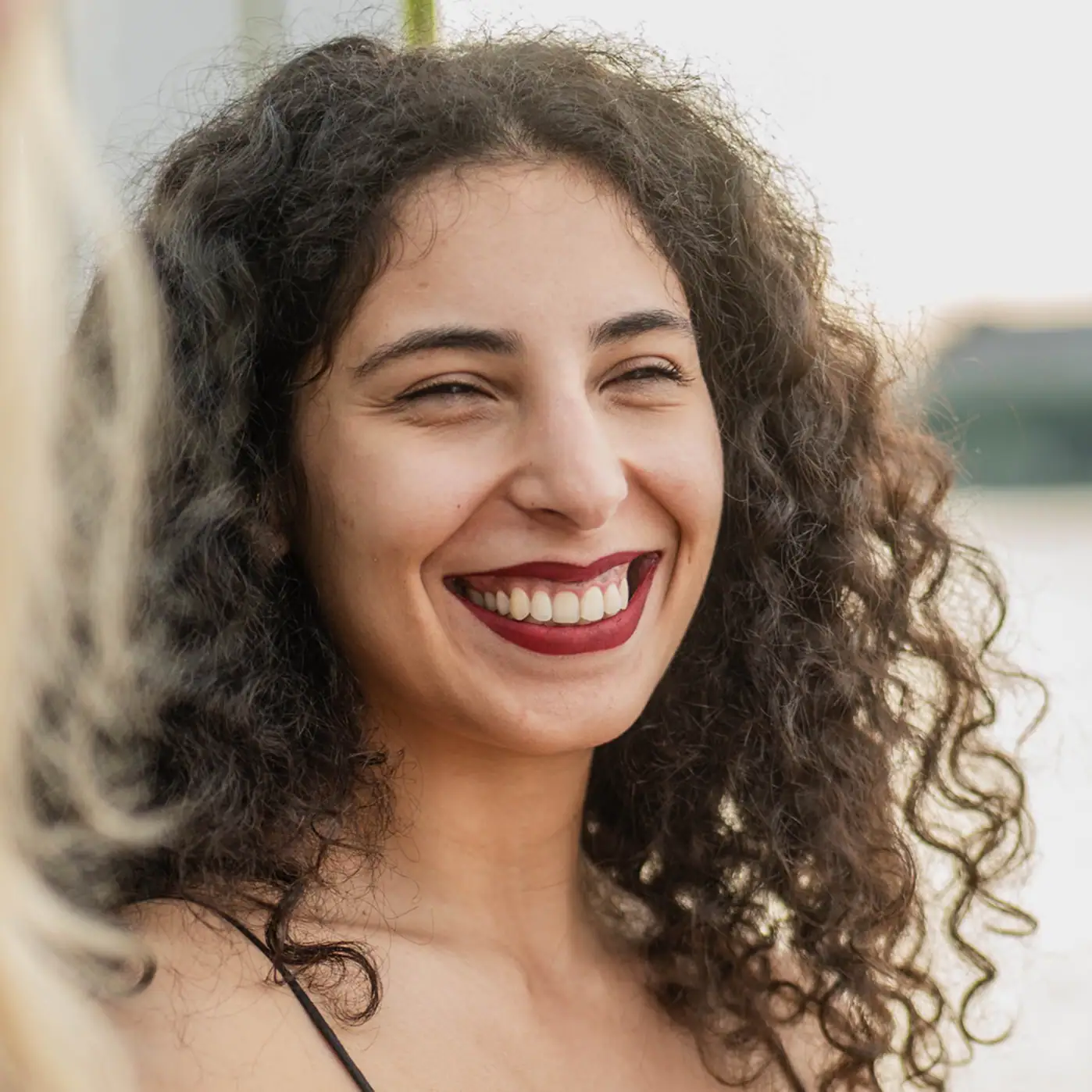 Jeune femme souriante aux cheveux bouclés et au rouge à lèvres rouge, dehors à la lumière du jour.