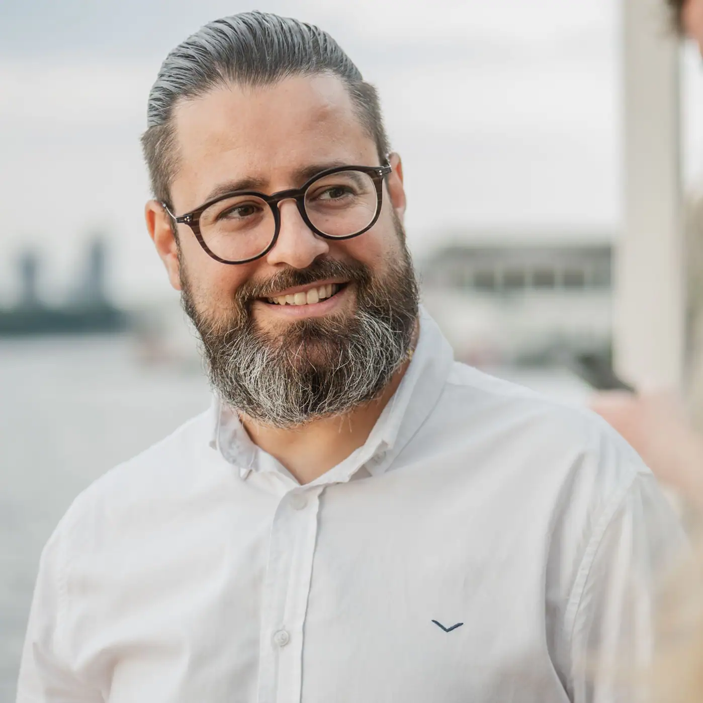 Hombre con gafas y barba sonríe mientras pasea junto al agua.