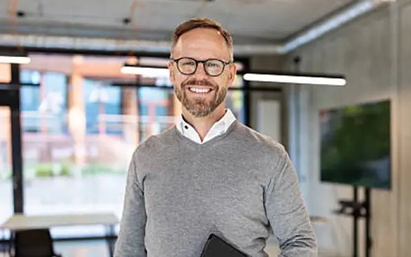 Ein Mann mit Brille und grauem Pullover lächelt in einem modernen Büro.