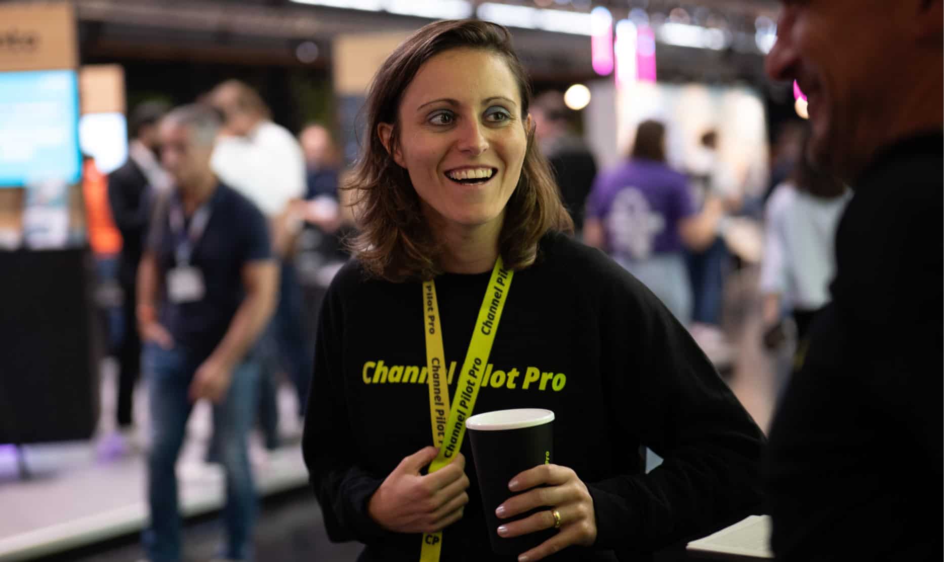 Freundliche Frau bei einer Messe im Gespräch, trägt ein "Chameleonot Pro" T-Shirt und hält einen Kaf.