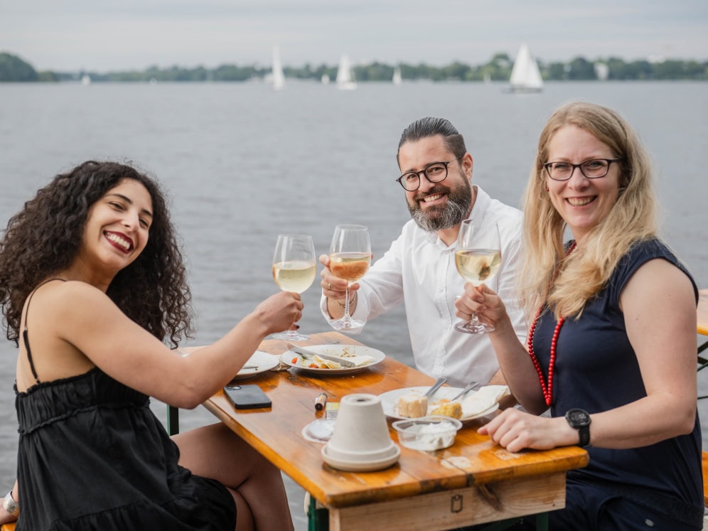 Ein fröhliches Trio beim Essen am Wasser, mit Wein und Blick auf Segelboote.