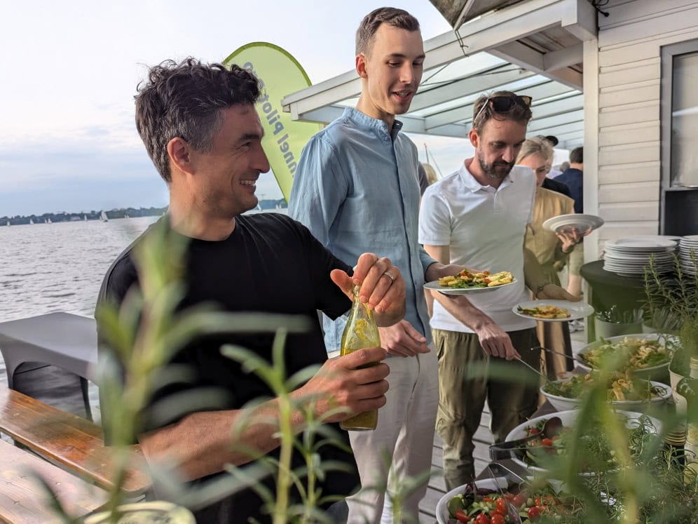 Fröhliche Menschen beim Buffet am Wasser, genießen ein gemeinsames Essen im Freien.